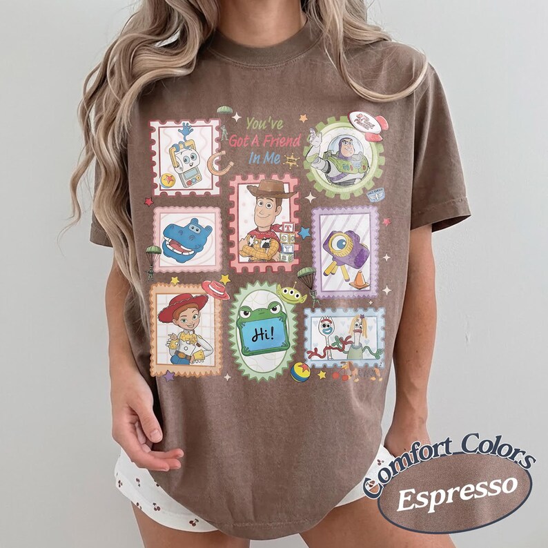 May include: A brown short-sleeve t-shirt featuring a graphic print of various Toy Story characters arranged in postage stamp-style frames. The text "You've Got A Friend In Me" is printed in the center, surrounded by illustrations of Woody, Buzz Lightyear, Jessie, and other characters.