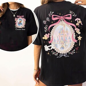 Pode incluir: Camiseta preta com um desenho do Pato Donald em uma moldura floral na frente. A parte de tr&aacute;s da camiseta exibe um desenho de castelo dentro de um oval emoldurado por fita rosa, cercado por elementos florais e do Mickey Mouse. O texto na frente diz "Custom Name".