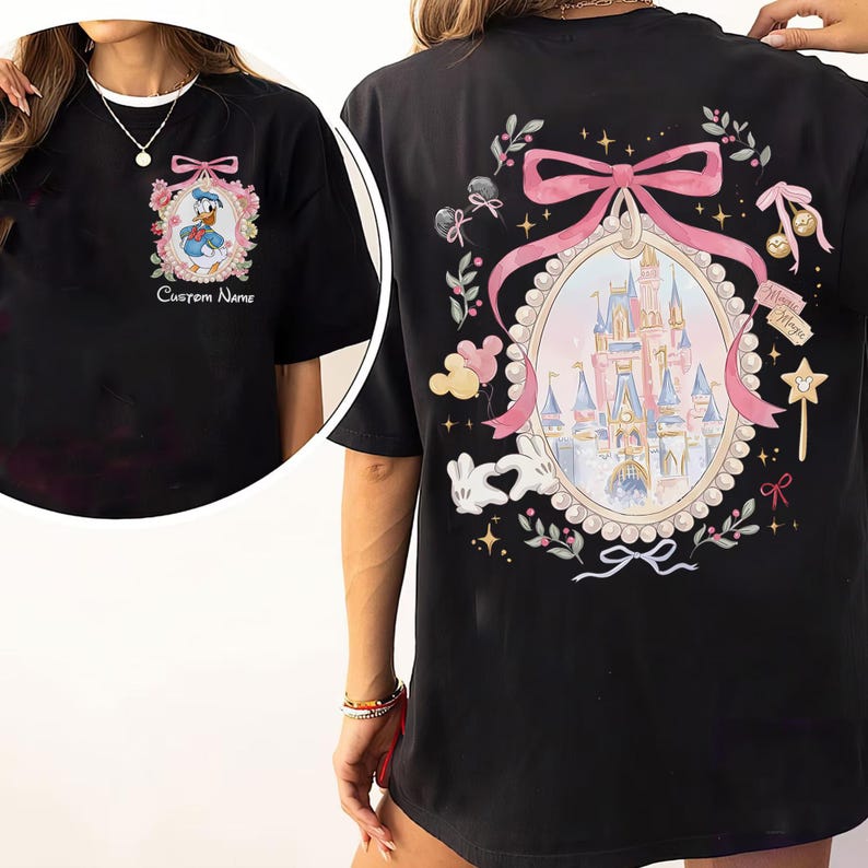 Pode incluir: Camiseta preta com um desenho do Pato Donald em uma moldura floral na frente. A parte de tr&aacute;s da camiseta exibe um desenho de castelo dentro de um oval emoldurado por fita rosa, cercado por elementos florais e do Mickey Mouse. O texto na frente diz "Custom Name".