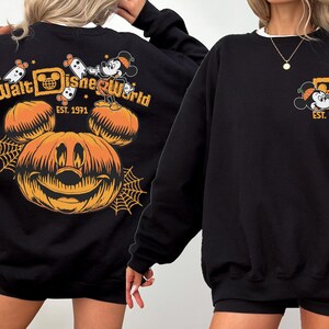 May include: Black sweatshirt featuring a Halloween-themed Walt Disney World design. The design includes a smiling jack-o'-lantern, Mickey Mouse, ghosts, and the text "Walt Disney World EST. 1971". The sweatshirt has a white collar.