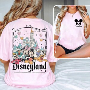 K&ouml;nnte beinhalten: Rosa T-Shirt mit einem Aufdruck von Mickey Mouse und seinen Freunden vor dem Disneyland-Schloss. Der Text "Disneyland - 1955" befindet sich unter dem Aufdruck.