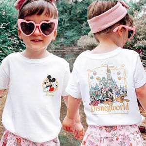 May include: Two children wearing white t-shirts with Disney World designs. One shirt features Mickey Mouse and the text "Disney". The other shirt features a drawing of Cinderella's castle and the text "Disneyworld 1971".