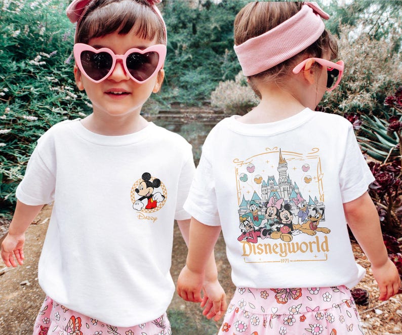 May include: Two children wearing white t-shirts with Disney World designs. One shirt features Mickey Mouse and the text "Disney". The other shirt features a drawing of Cinderella's castle and the text "Disneyworld 1971".