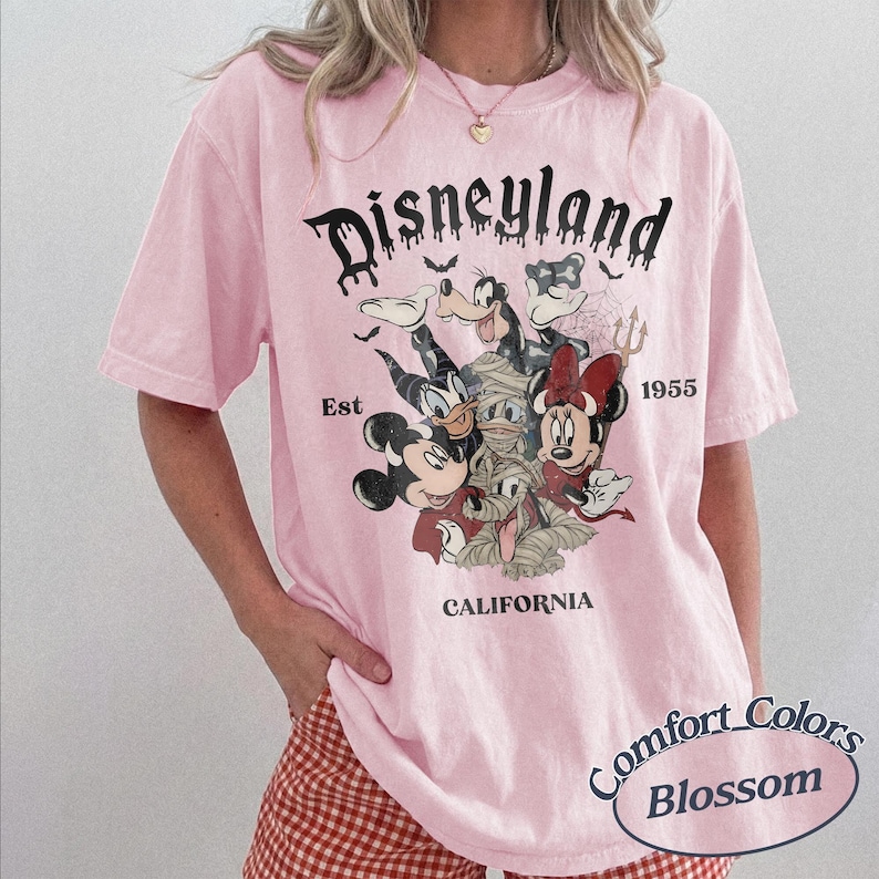 May include: A light pink short-sleeve t-shirt featuring a graphic print of Disney characters in Halloween costumes. The text reads Disneyland, Est 1955, and California. The design includes Mickey, Minnie, Donald, Goofy, and Pluto wrapped as a mummy.