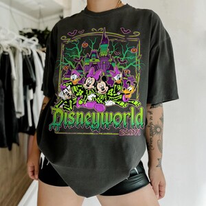 May include: Dark gray t-shirt featuring a Halloween-themed Disney World graphic. The design includes cartoon characters in skeleton costumes, a spooky castle, bats, and pumpkins. The text "Disneyworld Est. 1971" is also visible.