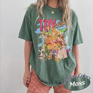May include: A moss green short-sleeve t-shirt featuring a graphic print of various animated characters and the text "TOY story". The shirt has a crew neckline and a relaxed fit, showcasing a colorful illustration of multiple figures in a group.