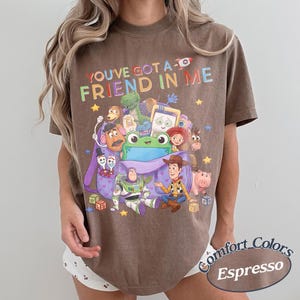 May include: A brown short-sleeve t-shirt featuring a graphic print of various Toy Story characters emerging from a purple backpack. The text "YOU'VE GOT A FRIEND IN ME" is printed in colorful letters above the characters, with small yellow stars.