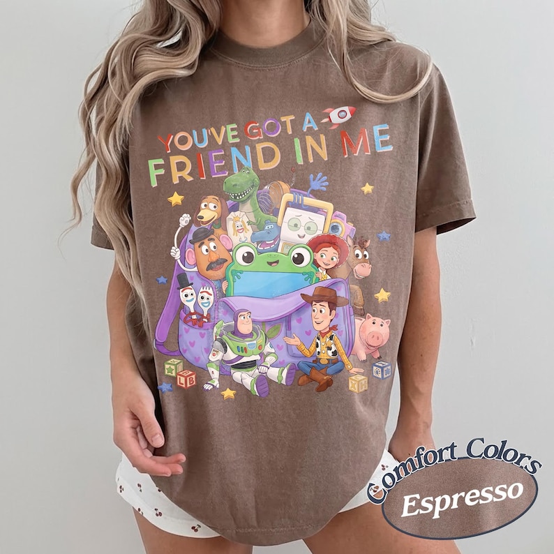 May include: A brown short-sleeve t-shirt featuring a graphic print of various Toy Story characters emerging from a purple backpack. The text "YOU'VE GOT A FRIEND IN ME" is printed in colorful letters above the characters, with small yellow stars.