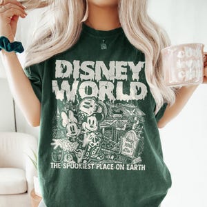 K&ouml;nnte beinhalten: Gr&uuml;nes T-Shirt mit einem abgenutzten wei&szlig;en Grafikdruck von Mickey Mouse und Minnie Mouse vor einer gruseligen Version von Disney World. Der Text "Disney World" befindet sich &uuml;ber den Figuren und "The Spookiest Place on Earth" darunter.
