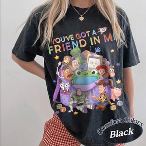 May include: A black acid-wash t-shirt featuring a colorful graphic of various animated toy characters emerging from a purple backpack. The text "YOU'VE GOT A FRIEND IN ME" is printed above the characters in multi-colored letters with a small rocket.