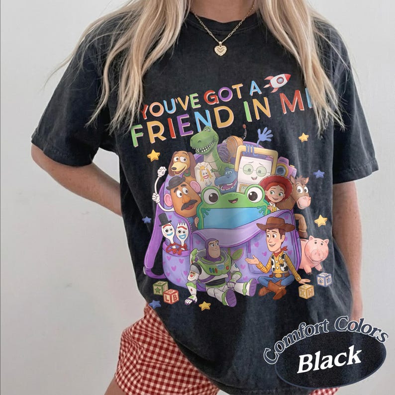 May include: A black acid-wash t-shirt featuring a colorful graphic of various animated toy characters emerging from a purple backpack. The text "YOU'VE GOT A FRIEND IN ME" is printed above the characters in multi-colored letters with a small rocket.