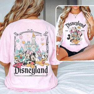 May include: Pink t-shirt with a graphic of Mickey Mouse and friends in front of the Disneyland castle. The text "Disneyland Est. 1955 California" is printed on the shirt.