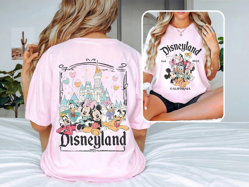 May include: Pink t-shirt with a graphic of Mickey Mouse and friends in front of the Disneyland castle. The text "Disneyland Est. 1955 California" is printed on the shirt.