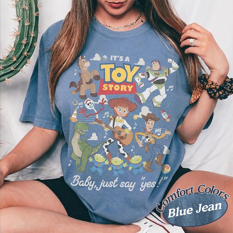 May include: A blue jean colored t-shirt featuring a Toy Story graphic with characters including Woody, Buzz Lightyear, Jessie, Rex, Bullseye, Forky, and the Aliens. The text reads IT'S A TOY STORY and Baby, just say yes! in white and yellow.