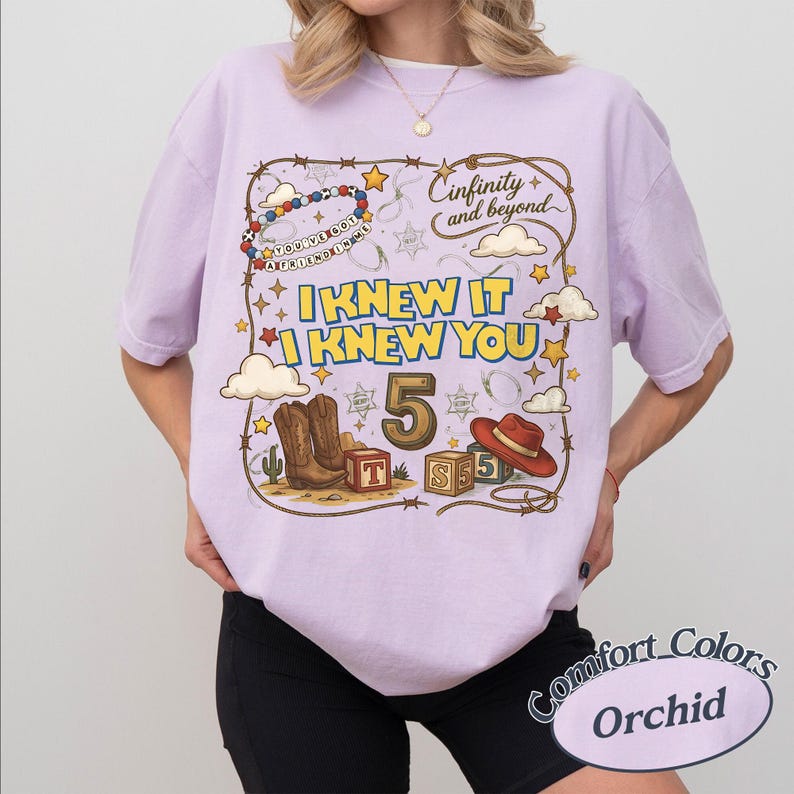 May include: A light purple t-shirt featuring a graphic print with a lasso border, cowboy boots, a red hat, wooden blocks with letters T, S, and 5, and the text "Cinfinity and beyond", "I KNEW IT I KNEW YOU", "5", and "YOU'VE GOT A FRIEND IN ME".