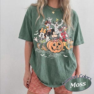 May include: A moss green short-sleeve t-shirt featuring a graphic print of Disney characters in Halloween costumes, including Mickey Mouse, Minnie Mouse, Donald Duck, Goofy, and Pluto around a jack-o'-lantern, with text signatures and bats.