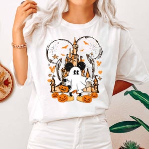 May include: White t-shirt with a Halloween design. The graphic features a ghost character, pumpkins, a castle, and Mickey Mouse ears. The design is in orange, black, and white.