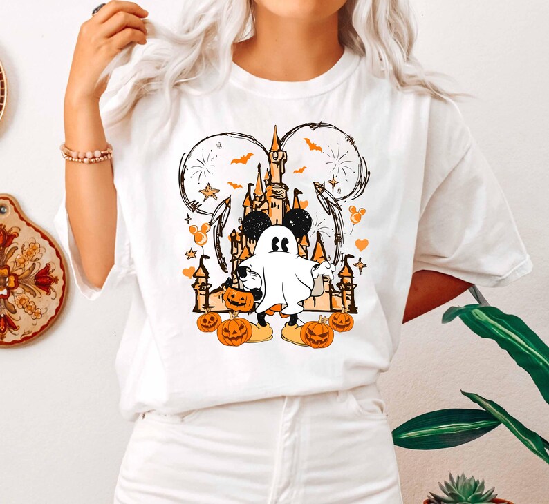 May include: White t-shirt with a Halloween design. The graphic features a ghost character, pumpkins, a castle, and Mickey Mouse ears. The design is in orange, black, and white.