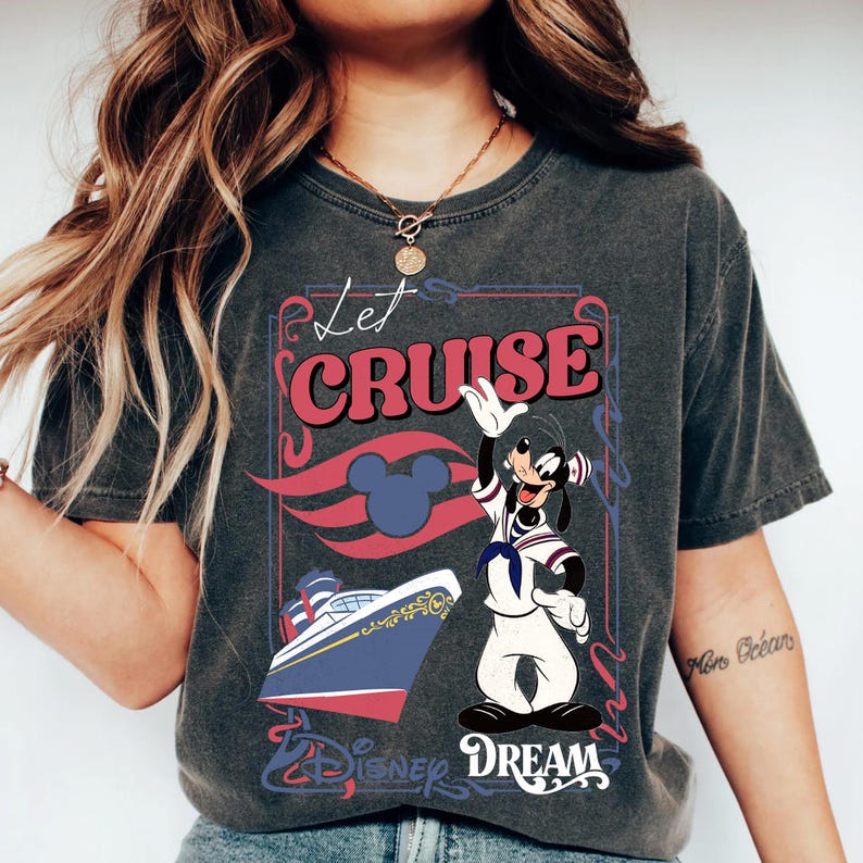 Może przedstawiać: Ciemnoszara koszulka z nadrukiem graficznym. Projekt zawiera słowa "Let Cruise" w kolorze czerwonym, statek wycieczkowy Disneya i machającą postacią z kresk&oacute;wki. Słowo "Dream" jest r&oacute;wnież obecne.