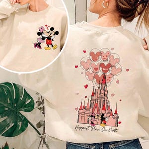 May include: Cream-colored sweatshirt featuring Mickey and Minnie Mouse. The front shows the couple embracing, while the back displays a castle with Mickey Mouse-shaped balloons and the text "Happiest Place On Earth."