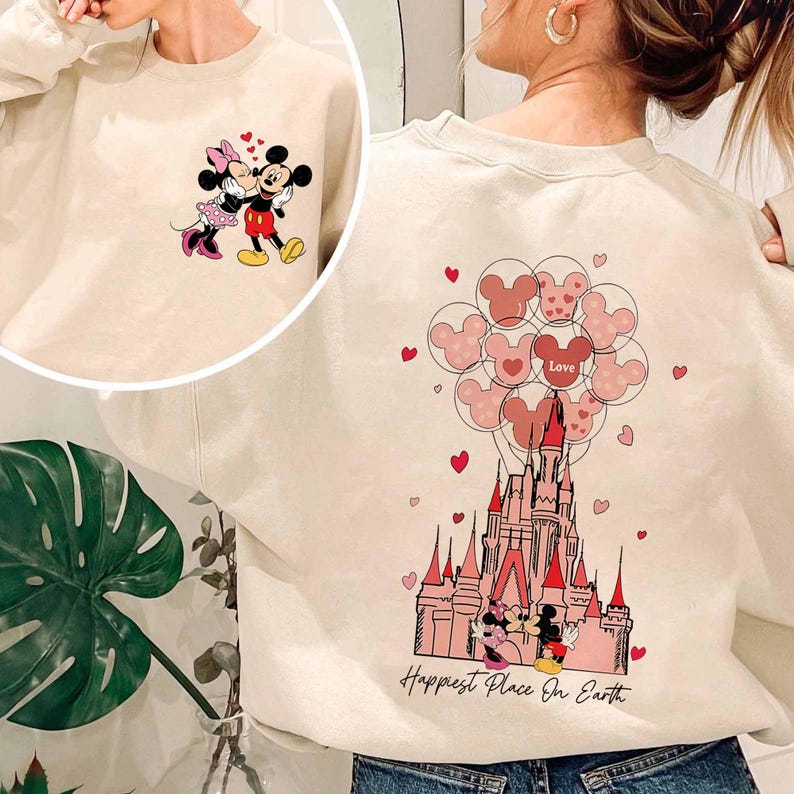 May include: Cream-colored sweatshirt featuring Mickey and Minnie Mouse. The front shows the couple embracing, while the back displays a castle with Mickey Mouse-shaped balloons and the text "Happiest Place On Earth."