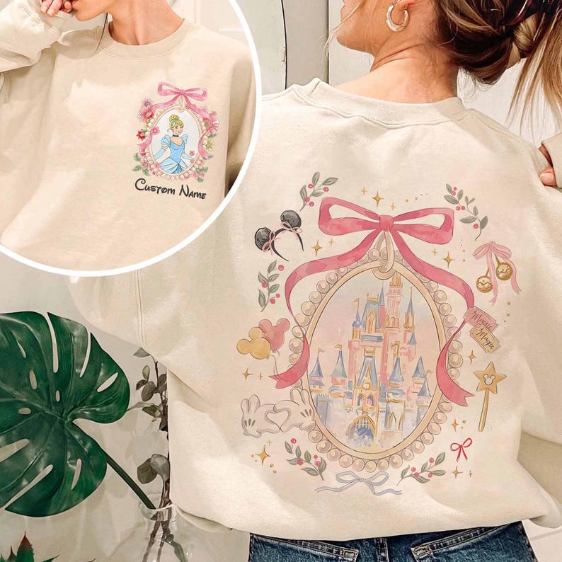 Pode incluir: Camisola creme com um design da Cinderela na frente e um design de castelo nas costas. O design frontal inclui o texto "Custom Name". O design traseiro inclui um castelo, orelhas da Minnie Mouse e uma varinha m&aacute;gica.