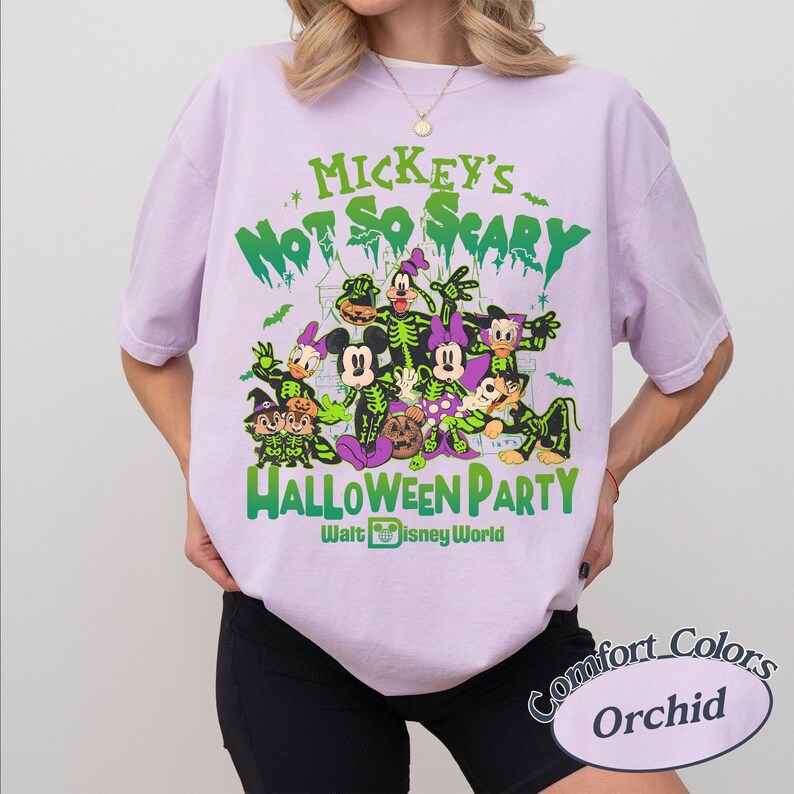 Op de afbeelding: Een lichtpaars T-shirt met een Halloween-thema Disney-afbeelding. Het ontwerp bevat verschillende personages in kostuums, met de tekst "Mickey's Not So Scary Halloween Party" in het groen. Comfort Colors merk, kleur Orchid.