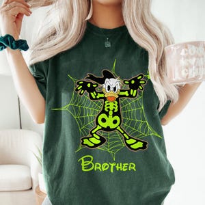 May include: Dark green t-shirt featuring a cartoon character in a skeleton costume, posed in front of a neon green spiderweb. The word "Brother" is printed in neon green at the bottom of the shirt.
