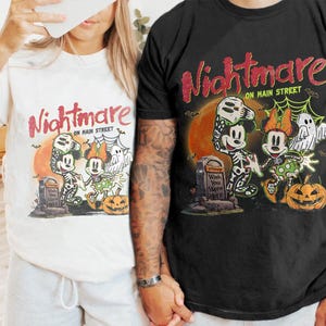 May include: White and black t-shirts with a Halloween-themed graphic. The design features Mickey and Minnie Mouse in skeleton costumes, a tombstone, a pumpkin, and the text "Nightmare on Main Street."
