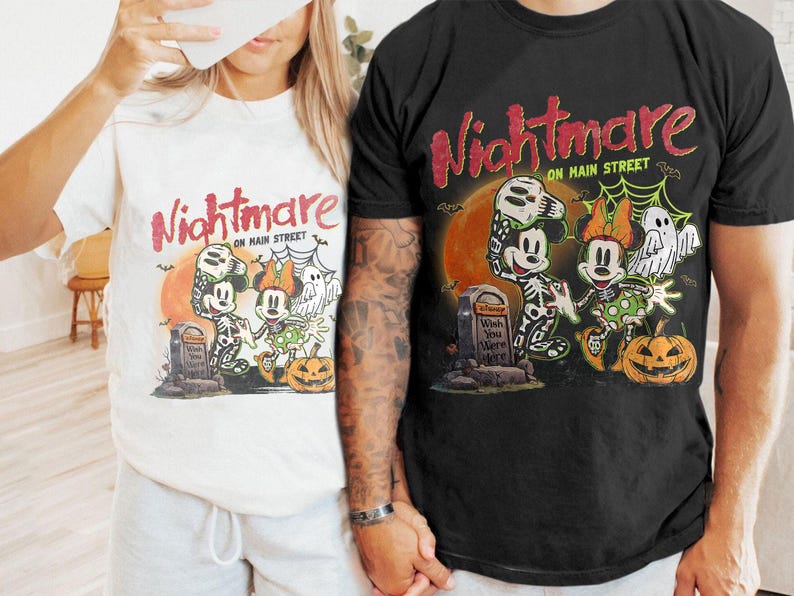 May include: White and black t-shirts with a Halloween-themed graphic. The design features Mickey and Minnie Mouse in skeleton costumes, a tombstone, a pumpkin, and the text "Nightmare on Main Street."