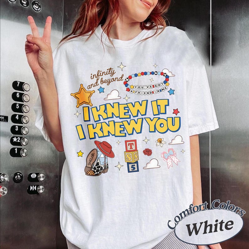 May include: A white t-shirt featuring colourful graphic prints including a yellow star, a beaded bracelet, a cowboy boot, and alphabet blocks. The text reads infinity and beyond, you've got a friend in me, and I KNEW IT I KNEW YOU.