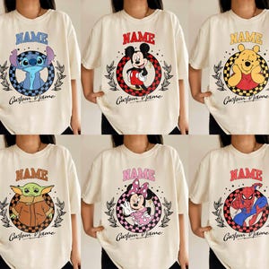 K&ouml;nnte beinhalten: Cremefarbene T-Shirts mit Cartoonfiguren: Stitch, Mickey Mouse, Winnie the Pooh, Grogu, Minnie Mouse und Spider-Man. Jedes Design enth&auml;lt das Wort "NAME" und "Custom Name".