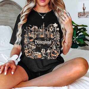 May include: Black t-shirt featuring a Disneyland design in orange and white. The graphic includes a castle, Mickey Mouse, pumpkins, and other park icons. The word "Disneyland" is printed in a stylized font.