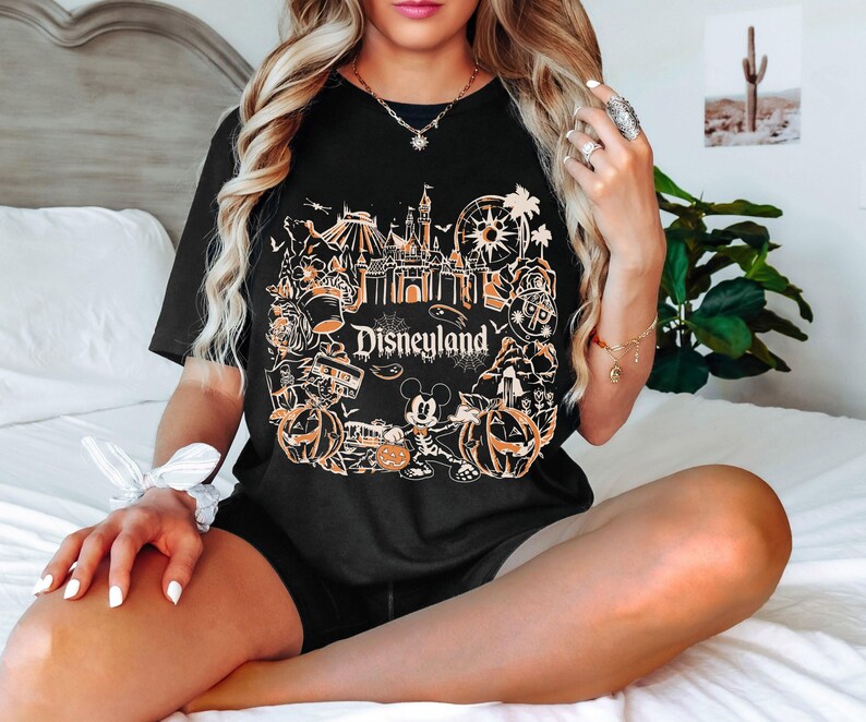 May include: Black t-shirt featuring a Disneyland design in orange and white. The graphic includes a castle, Mickey Mouse, pumpkins, and other park icons. The word "Disneyland" is printed in a stylized font.