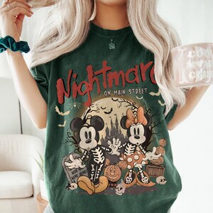 May include: Dark green t-shirt with a Halloween-themed graphic. The design features Mickey and Minnie Mouse dressed as skeletons in front of a full moon, with the words "Nightmare on Main Street" in red.