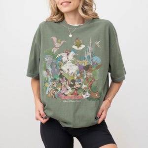 K&ouml;nnte beinhalten: Olivgr&uuml;nes T-Shirt mit einem farbenfrohen Aufdruck von Disney-Figuren, darunter Mickey Mouse, Dumbo und Peter Pan. Der Text "Magic Kingdom, Walt Disney World" ist unten auf dem Design aufgedruckt.