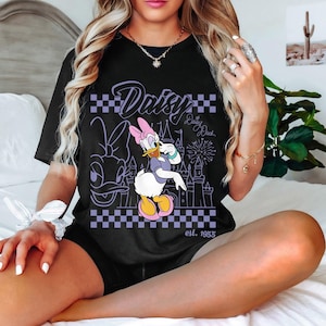 May include: Black t-shirt featuring a graphic of Daisy Duck in a purple and white checkered design. The design includes the word "Daisy" and "Daisy Duck" with a castle and fireworks. The text "est. 1955" is also visible.