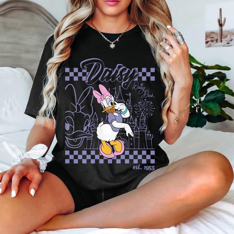 May include: Black t-shirt featuring a graphic of Daisy Duck in a purple and white checkered design. The design includes the word "Daisy" and "Daisy Duck" with a castle and fireworks. The text "est. 1955" is also visible.