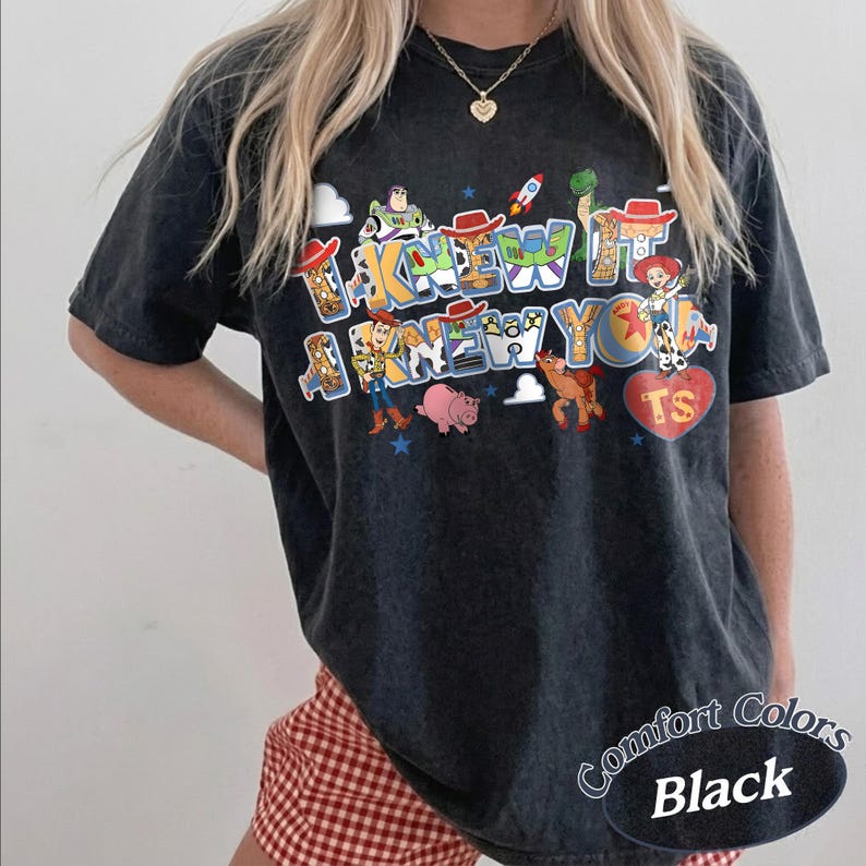 May include: A black short-sleeve t-shirt featuring a graphic print of Toy Story characters including Woody, Buzz Lightyear, Jessie, Rex, Bullseye, and Hamm. The text reads I KNEW IT I KNEW YOU with a red heart shape containing the letters TS.
