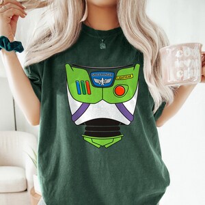 May include: Dark green t-shirt featuring a graphic of a Buzz Lightyear costume. The design includes a green helmet, white chest, and purple accents. The text "SPACE RANGER" and "LIGHTYEAR" are visible on the design.