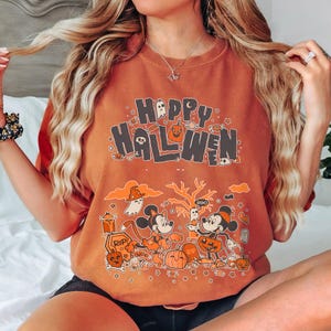 Puede incluir: Camiseta naranja con un gr&aacute;fico de Halloween. La camiseta presenta las palabras "Happy Halloween" sobre una ilustraci&oacute;n de dibujos animados de Mickey Mouse y Minnie Mouse disfrazados con disfraces de Halloween. Tambi&eacute;n se incluyen calabazas, fantasmas y un &aacute;rbol espeluznante.