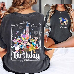 May include: Dark gray t-shirt featuring a colorful graphic of Disney characters, a castle, balloons, and fireworks. The text "Birthday Squad" is displayed in a white, decorative font. The shirt has a vintage, washed look.