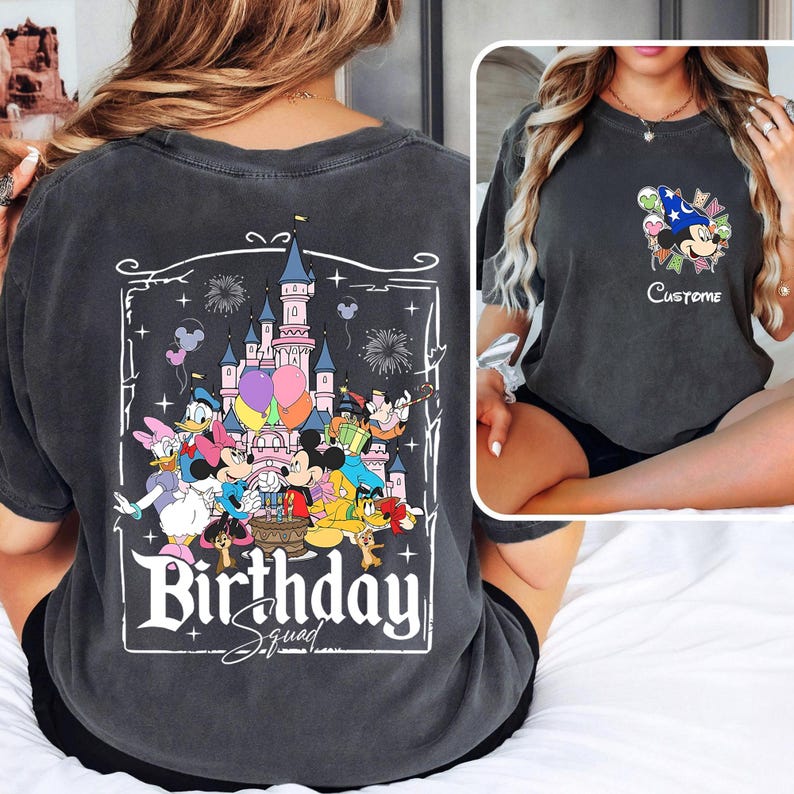 May include: Dark gray t-shirt featuring a colorful graphic of Disney characters, a castle, balloons, and fireworks. The text "Birthday Squad" is displayed in a white, decorative font. The shirt has a vintage, washed look.