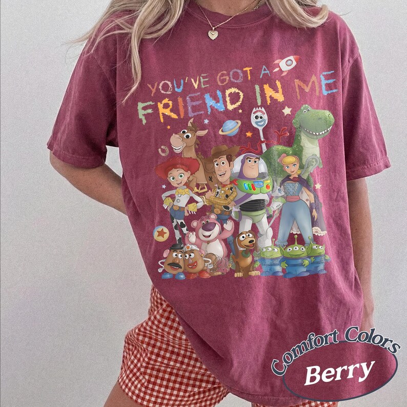 May include: A berry-colored short-sleeve t-shirt featuring a graphic print of various animated characters from the Toy Story franchise. The text "YOU'VE GOT A FRIEND IN ME" is printed in colorful letters above the group of characters.