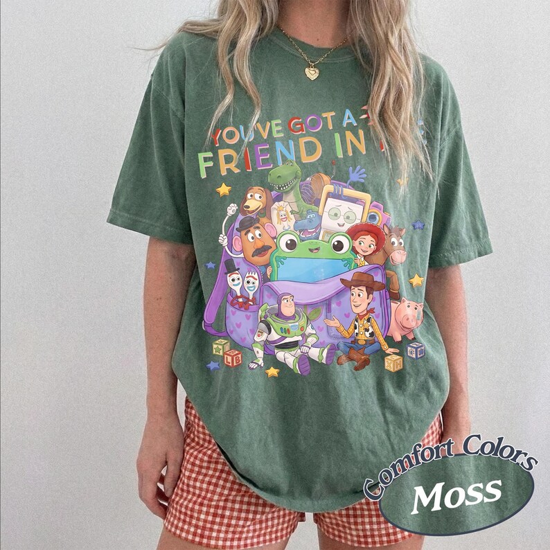 May include: A moss green short-sleeve t-shirt featuring a colorful graphic print of various animated characters and a purple backpack. The text "YOU'VE GOT A FRIEND IN ME" is printed in multi-colored letters above the character illustration.