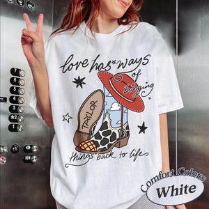 May include: A white t-shirt featuring a graphic of a cowboy boot with a cow print pattern, a red hat, and the text "love has ways of bringing things back to life" along with the name "TAYLOR" and the number "13" printed on the boot design.