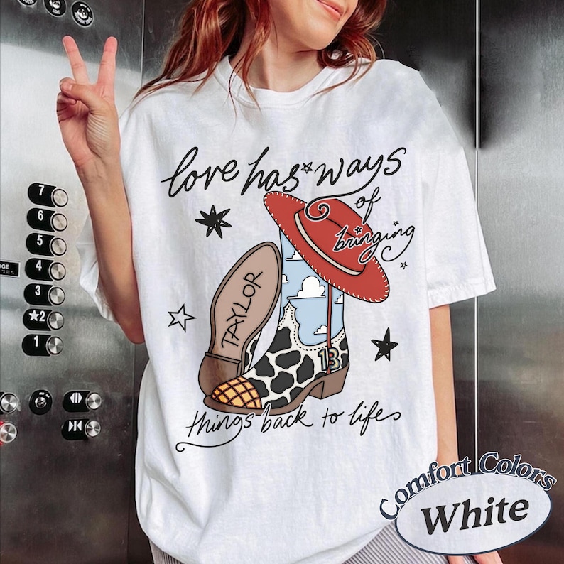 May include: A white t-shirt featuring a graphic of a cowboy boot with a cow print pattern, a red hat, and the text "love has ways of bringing things back to life" along with the name "TAYLOR" and the number "13" printed on the boot design.