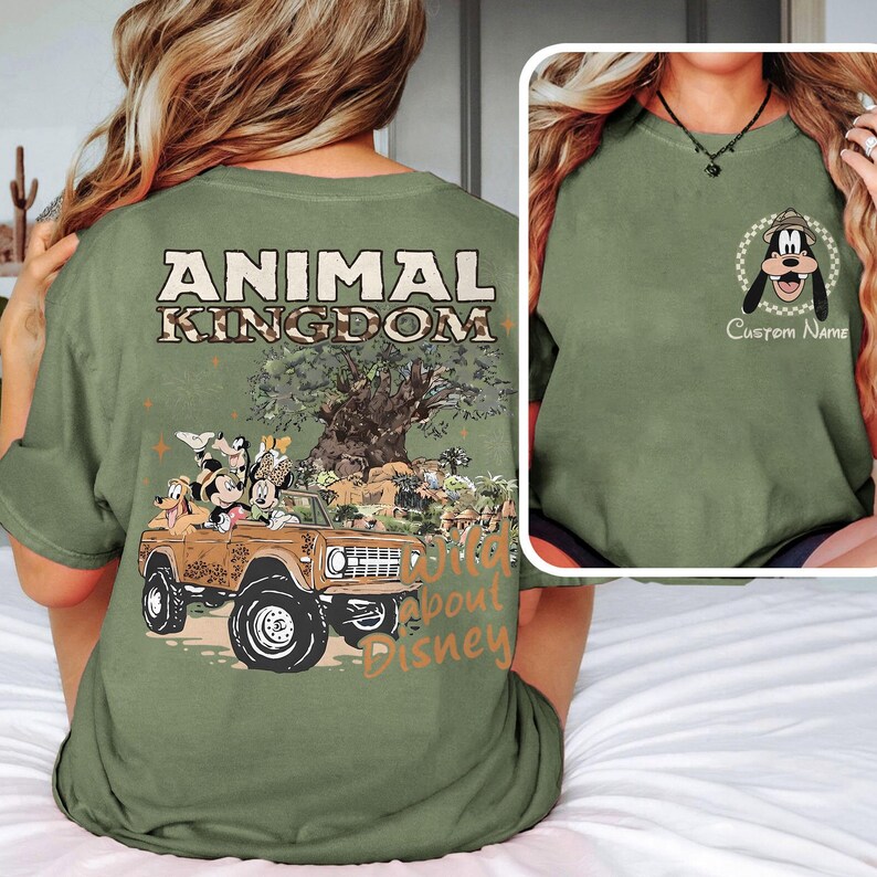 May include: Olive green t-shirt featuring a graphic of Disney characters in a brown jeep with the text "ANIMAL KINGDOM" and "Wild About Disney." The shirt also has a smaller image of Goofy with the text "Custom Name."