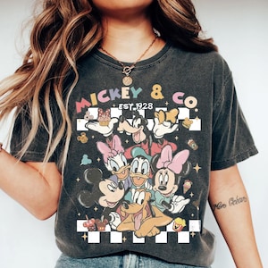 May include: Dark gray t-shirt featuring a colorful graphic of Mickey Mouse, Minnie Mouse, Donald Duck, Daisy Duck, Goofy, and Pluto. The shirt has the text "Mickey & Co. Est. 1928" in a retro font, with a checkered background.