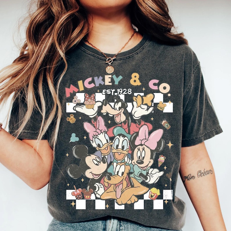 May include: Dark gray t-shirt featuring a colorful graphic of Mickey Mouse, Minnie Mouse, Donald Duck, Daisy Duck, Goofy, and Pluto. The shirt has the text "Mickey & Co. Est. 1928" in a retro font, with a checkered background.